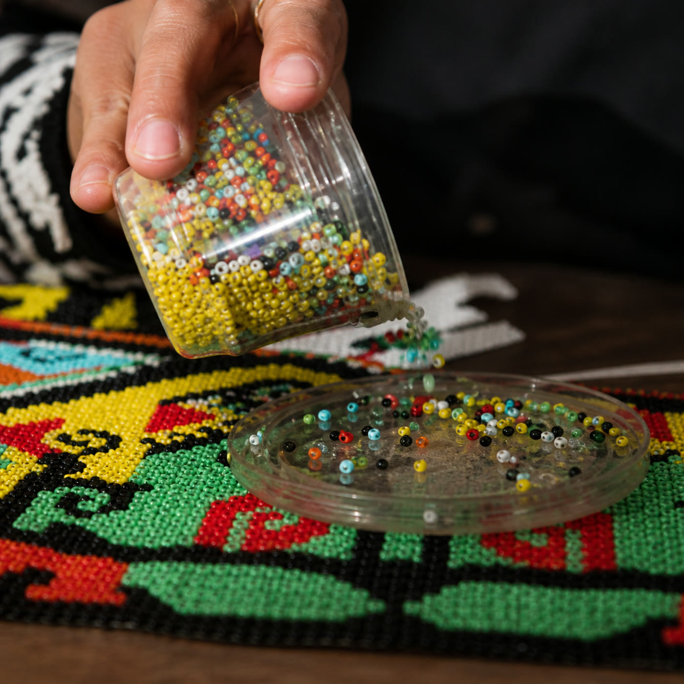 A hand pouring multicoloured glass seed beads into a sorting tray over finished beadwork
